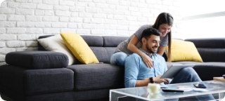 Man smiles while sitting on the floor looking at a tablet, woman sits behind him on a couch smiling.